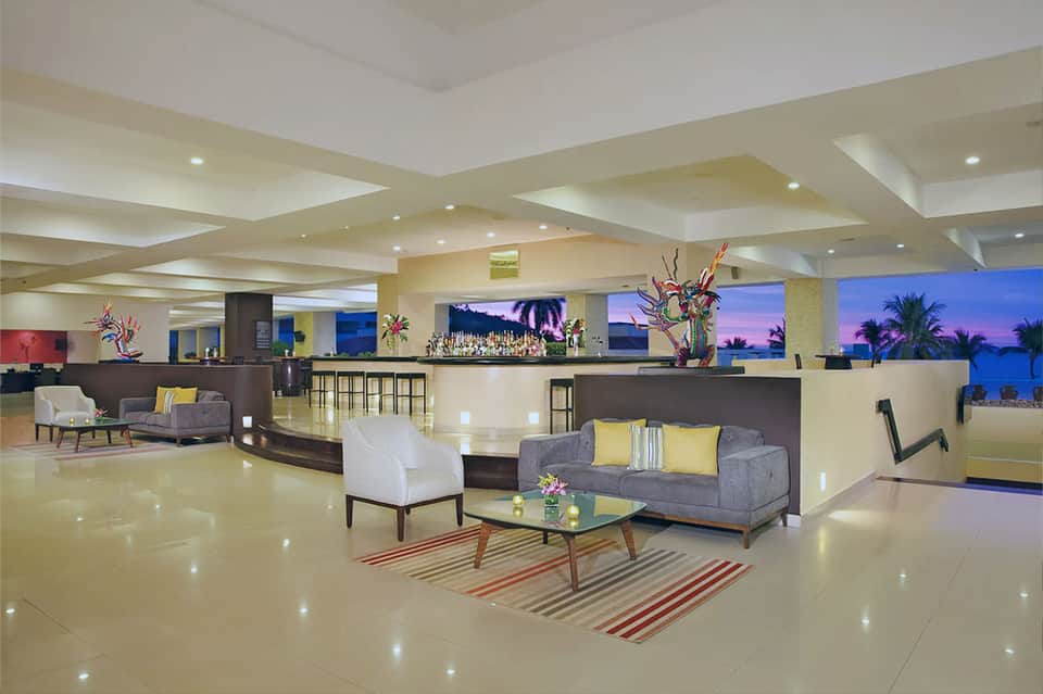 Spacious hotel lobby with beige and gray seating, bar area, and ocean view windows at evening