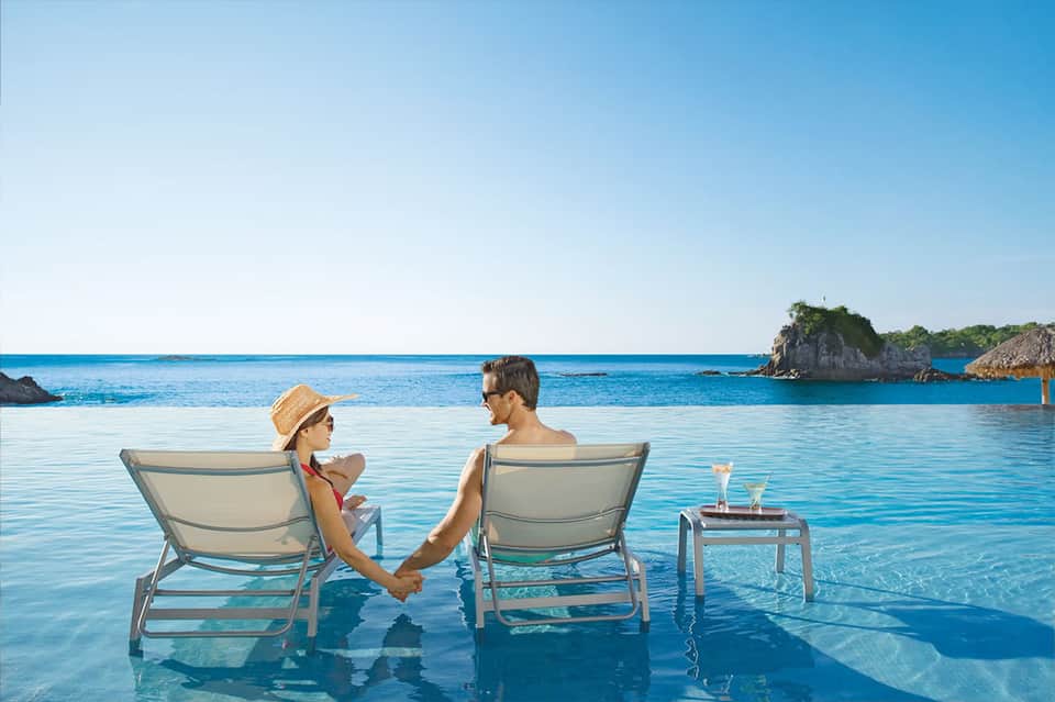 Couple relaxing in lounge chairs in shallow turquoise water with tropical islands visible