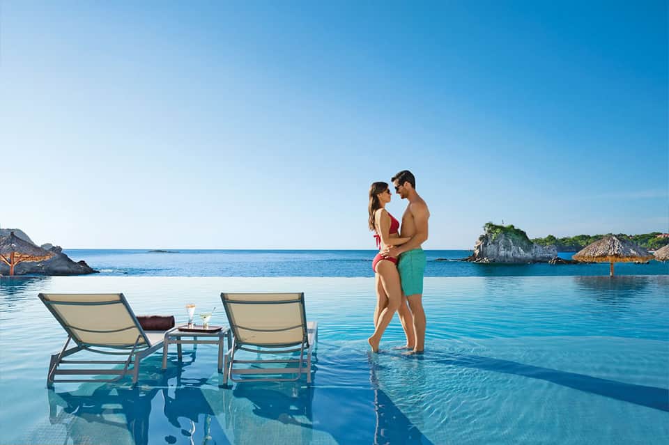 Couple wading in infinity pool overlooking calm blue ocean with rocky islands