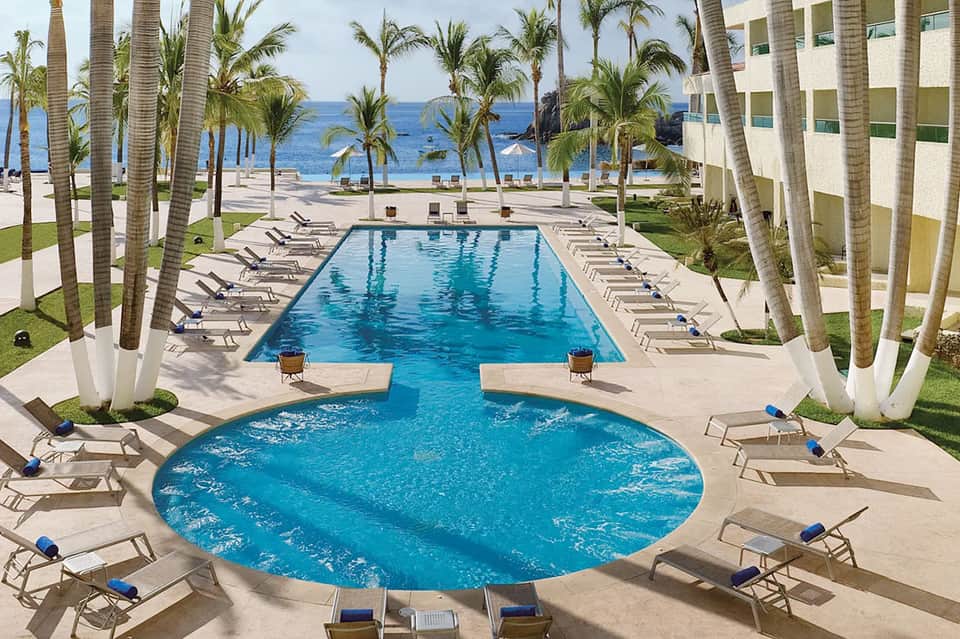 Beachfront resort pool with palm trees, lounge chairs, ocean view, and modern architecture