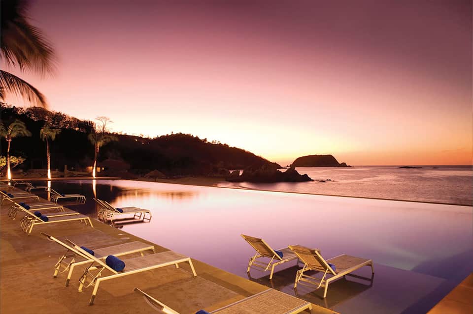 Beach lounge chairs at twilight with calm ocean, palm trees, and rocky shoreline