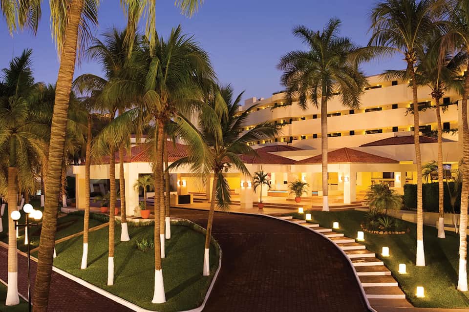 Resort exterior at dusk with illuminated building, palm trees, and manicured lawn