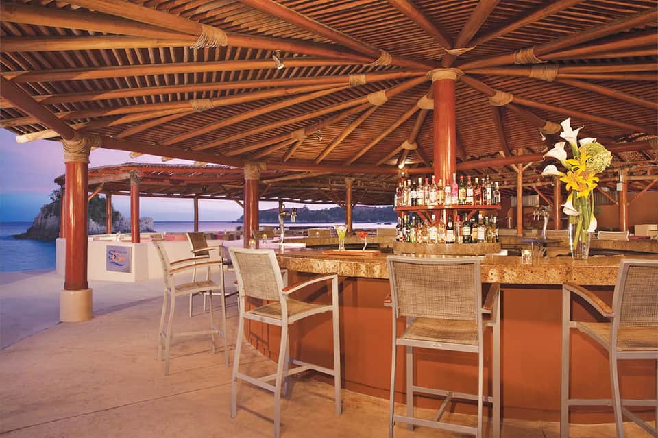 Beach bar at sunset with wooden pergola, tiki torches, ocean view, tropical chairs, waterfront setting