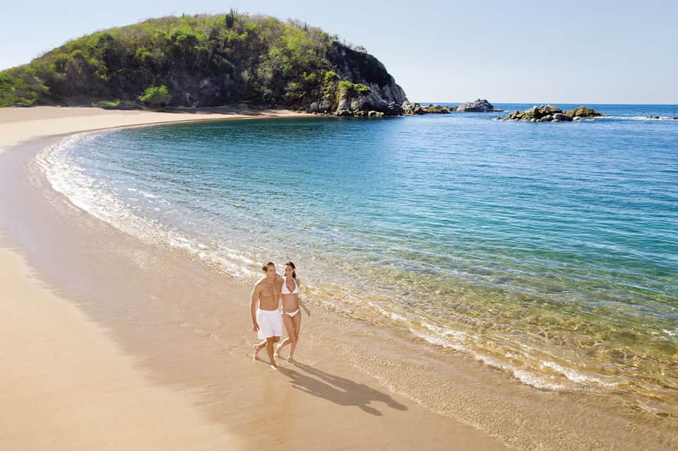 Couple wading in turquoise ocean water near pristine white sand beach with rocky headland