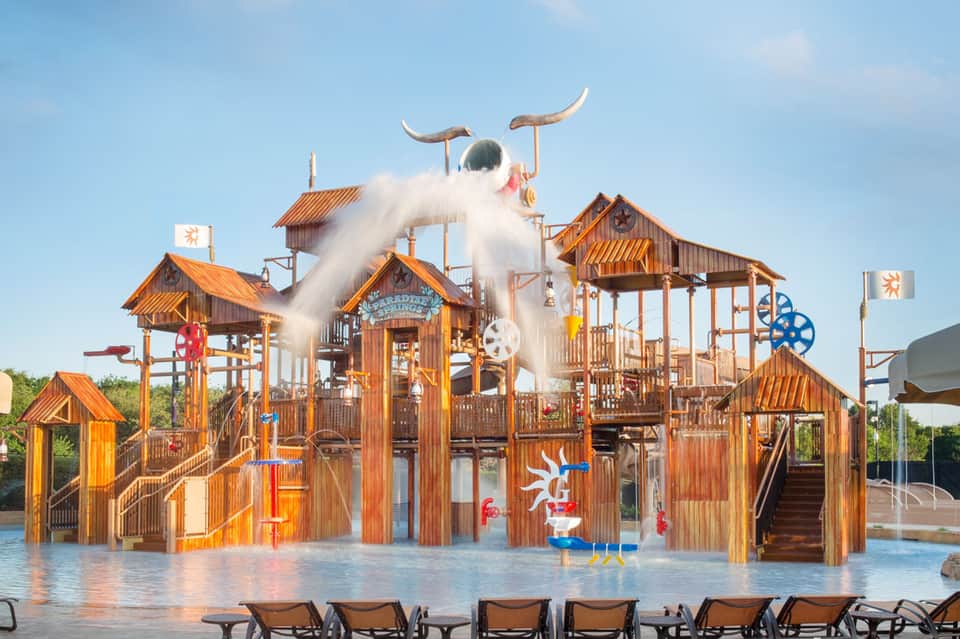 Wooden water park structure with white water slide and bull horns, surrounded by shallow pool and lounge chairs