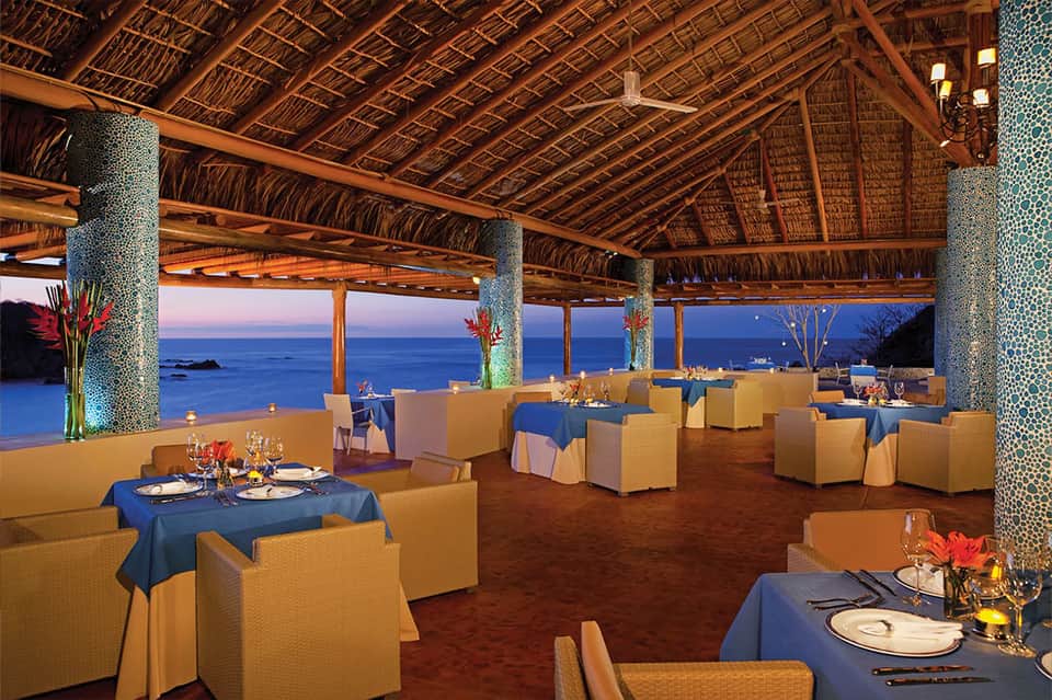 Fine dining restaurant with thatched roof, ocean views, and elegantly set tables