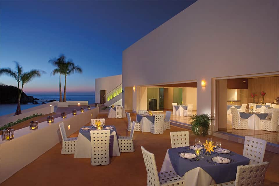 Beachfront restaurant terrace with dining tables at dusk, palm trees and ocean beyond