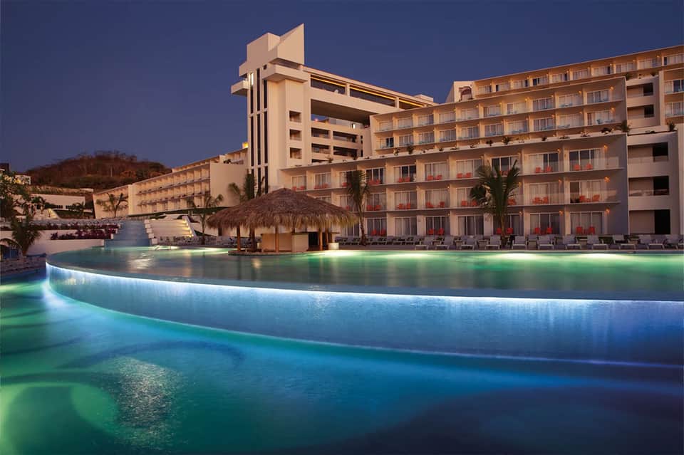 Curved illuminated pool at dusk with hotel building and thatched bar structure at night