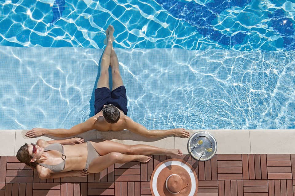 Man floating in crystal-clear pool while woman relaxes poolside with hat and refreshment