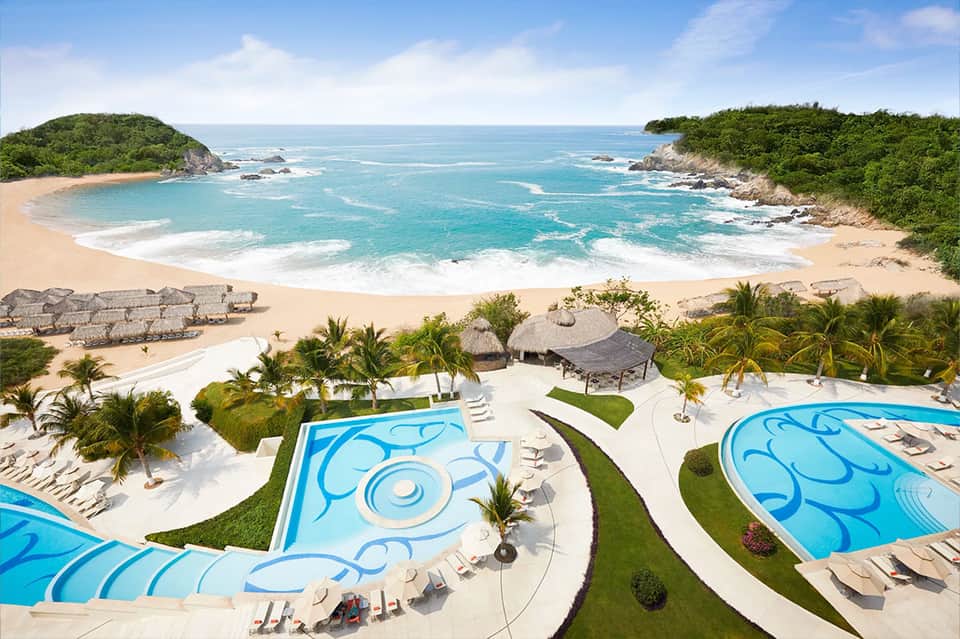 Aerial view of curved resort pools overlooking a secluded cove beach with turquoise waters