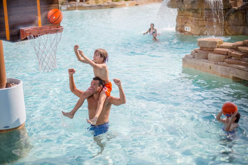 Family playing basketball in a resort pool with water features and rock formations
