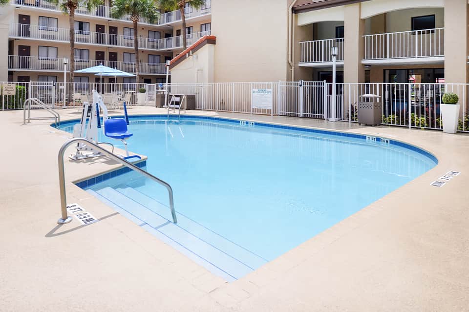 Small rectangular pool with safety railings, slide, lounge chairs, and multi-story residential building behind