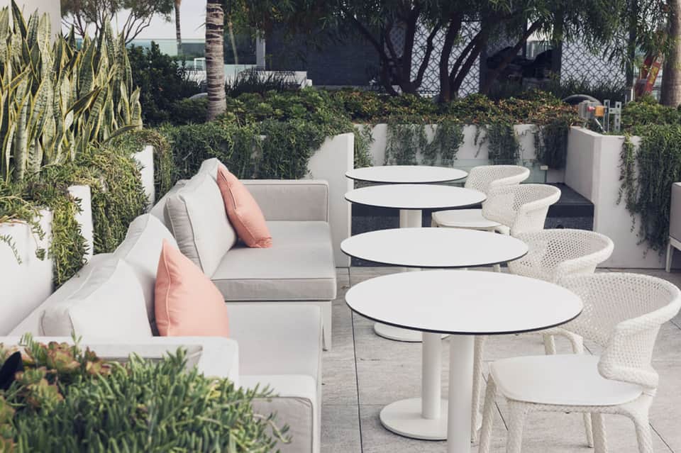 Modern outdoor terrace with white lounge seating, round tables, and lush green plants