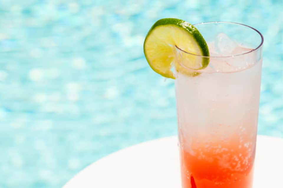 Coral-colored cocktail with lime slice garnish on white pool deck with turquoise water backdrop