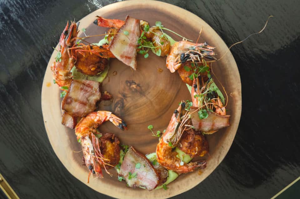 Overhead view of grilled shrimp and bacon appetizers arranged on a round wooden plate