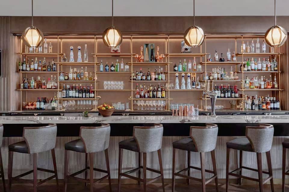 Modern bar counter with extensive liquor bottle display on wooden shelving, pendant lighting, and upholstered seating