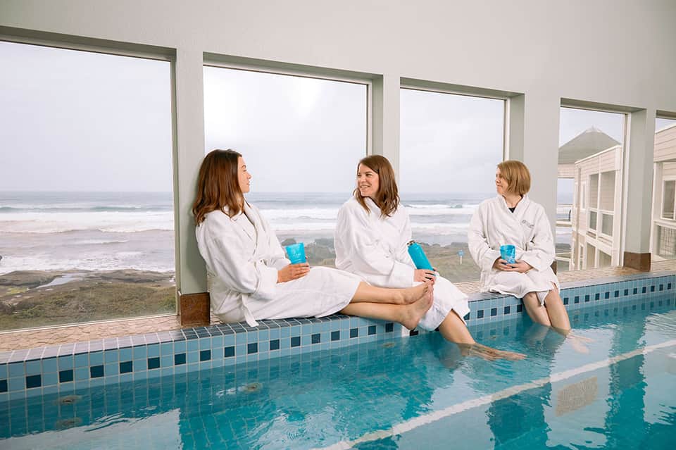 Three women in white robes sitting poolside with ocean beach view through large windows