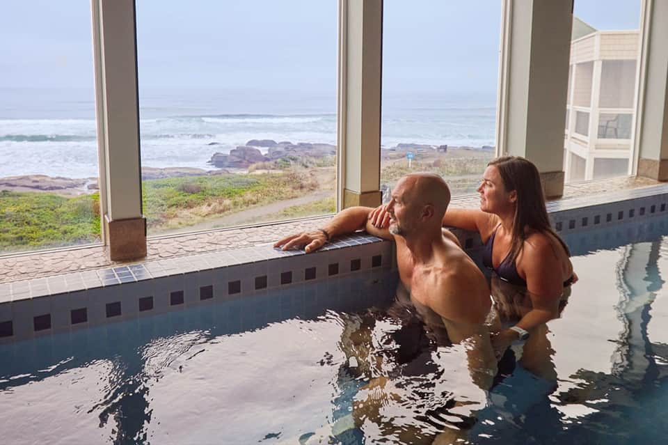 Couple relaxing in an indoor thermal pool with ocean and rocky coastline views through windows
