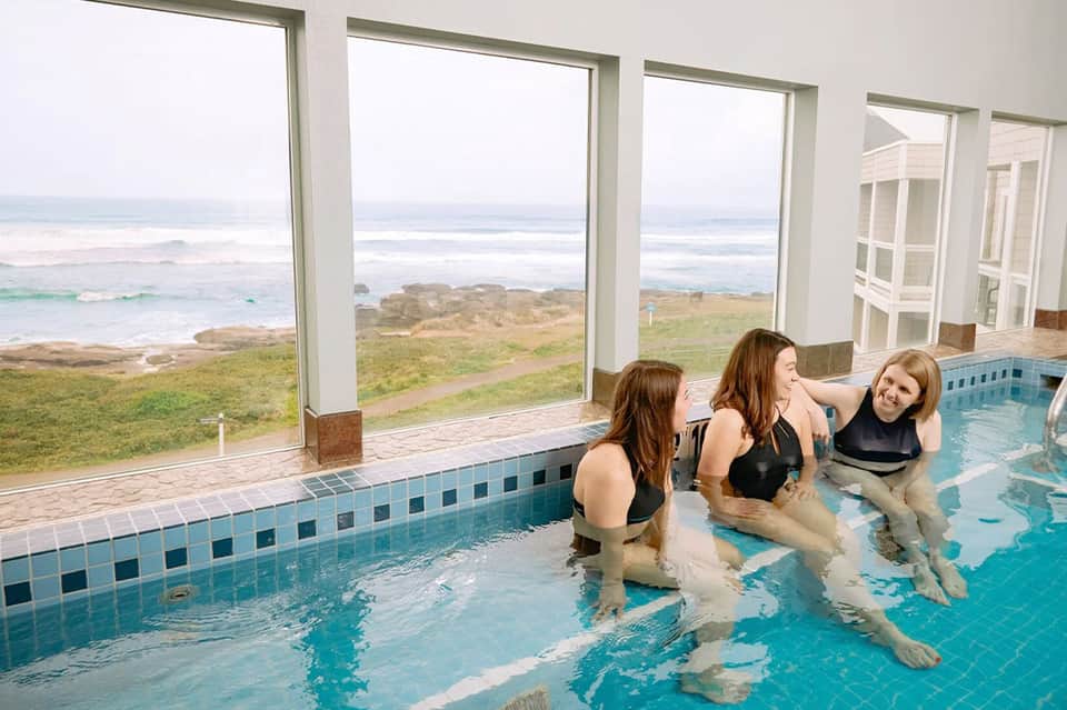 Three women in indoor thermal pool with ocean and coastal views through large windows