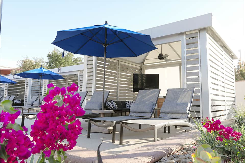 Modern poolside cabanas with blue umbrellas, lounge chairs, and pink flowering plants