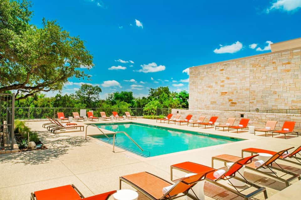 Modern pool area with bright orange lounge chairs, stone building, green trees, and clear blue sky.