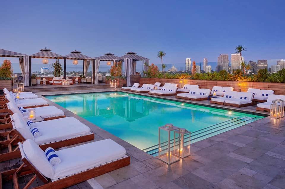Elevated rooftop pool at dusk with lounge beds, city skyline, and ambient lighting