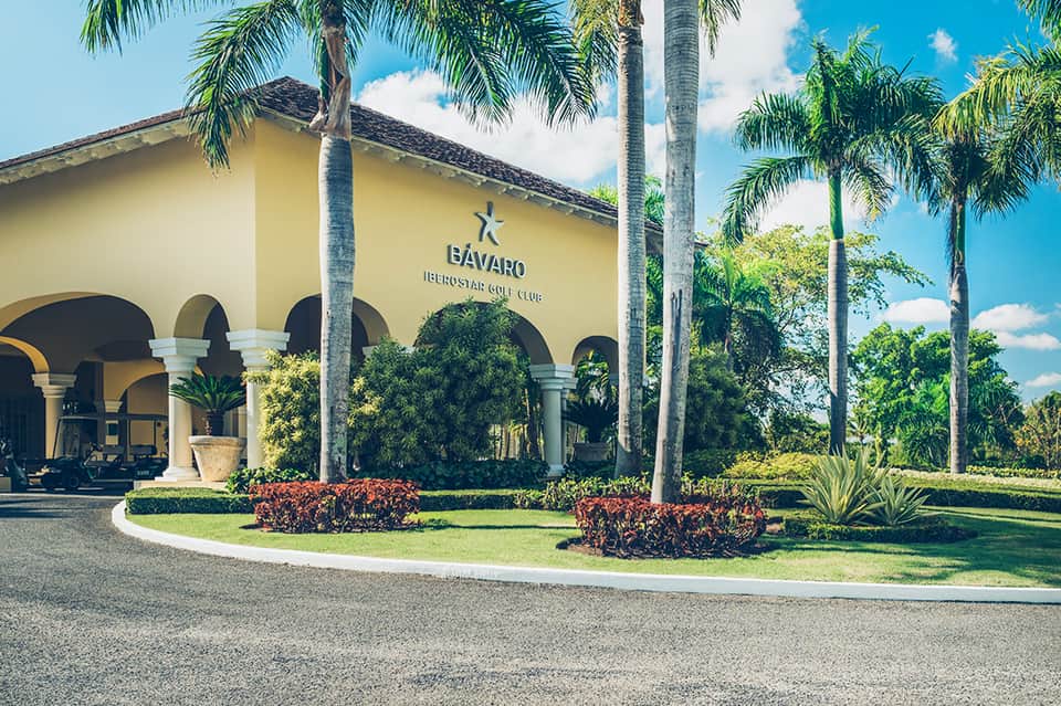 Yellow Spanish-style golf club building with white columns and palm tree-lined entrance drive