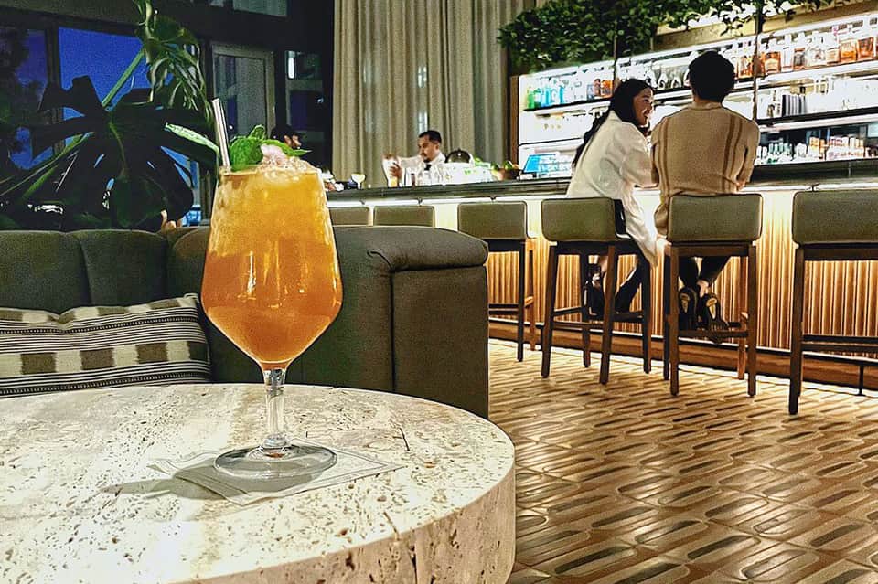 Modern bar lounge with green seating, tropical plants, and ambient lighting in evening atmosphere