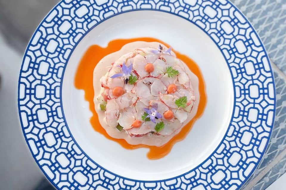Plated ceviche dish with sliced fish, orange sauce, and colorful edible flowers on decorative plate