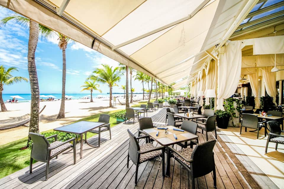 Beachfront dining area with cream sail shade, wooden deck, palms, and ocean horizon