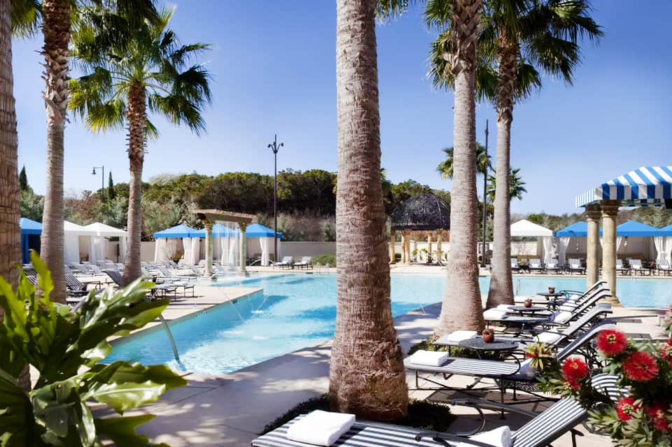 Large swimming pool with tall palm trees, lounge chairs, and covered cabanas