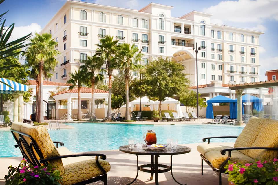 Resort pool with cream building, palm trees, white umbrellas, and lounge seating