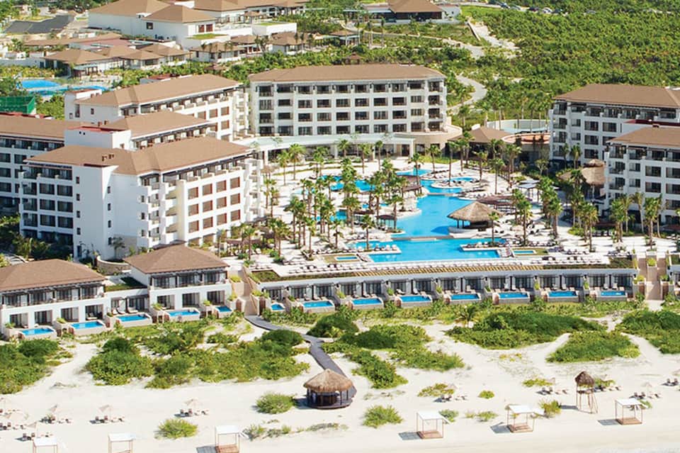 Aerial view of beachfront resort with multiple pools, white buildings, and surrounding palm trees