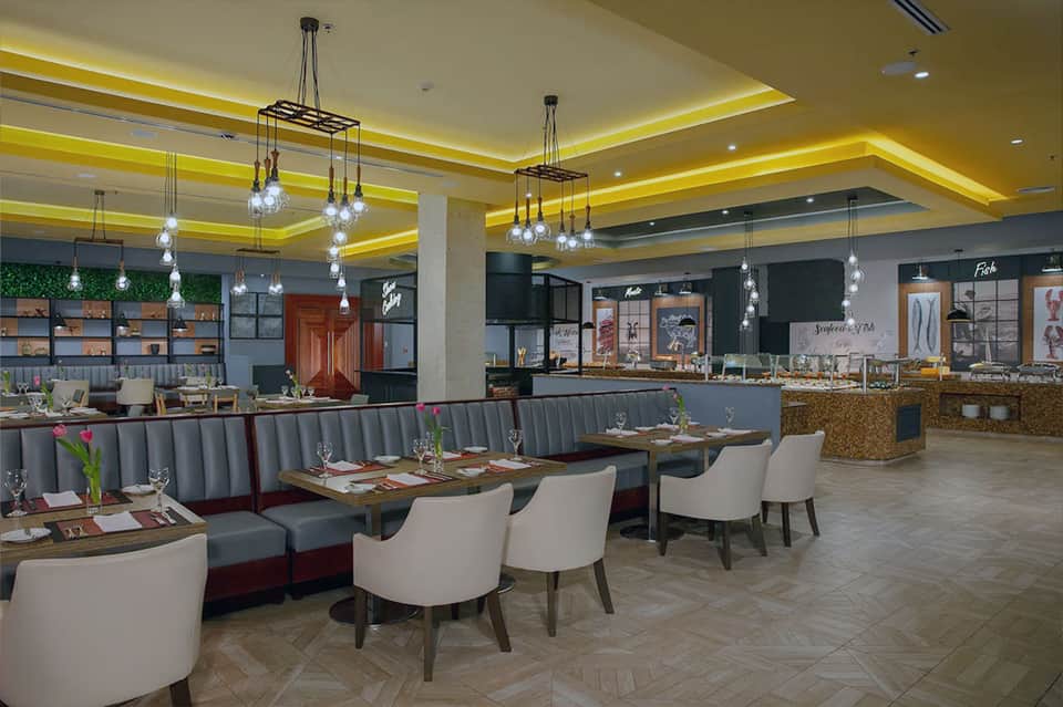 Restaurant dining area with yellow ceiling accent, hanging lights, gray banquettes, and modern furnishings