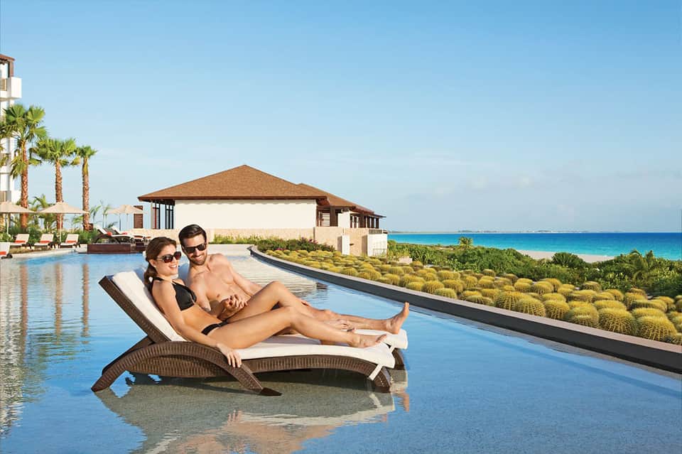 Couple relaxing on chaise lounge in infinity pool overlooking pristine beach and ocean