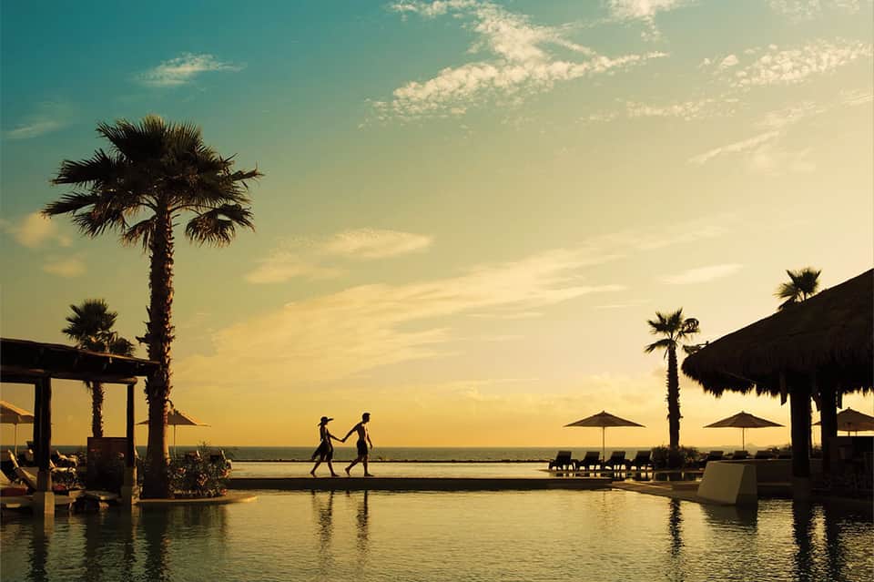 Couple walking on infinity pool deck at golden sunset with palm trees and thatched umbrellas