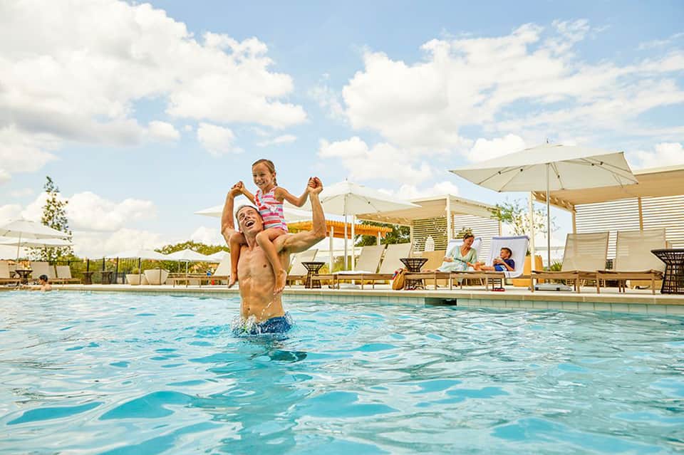 Father lifting child in resort pool with umbrellas and lounge seating in background