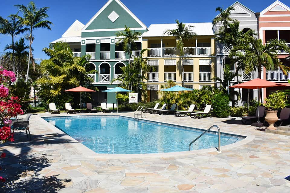Colorful colonial-style beachfront buildings with pool, lounge chairs, umbrellas, palm trees, and flowers
