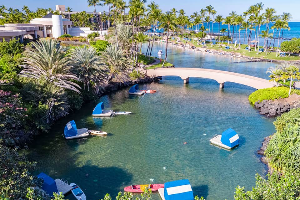 Aerial view of resort with lagoon-style pool, palm trees, boats, and blue ocean water in background
