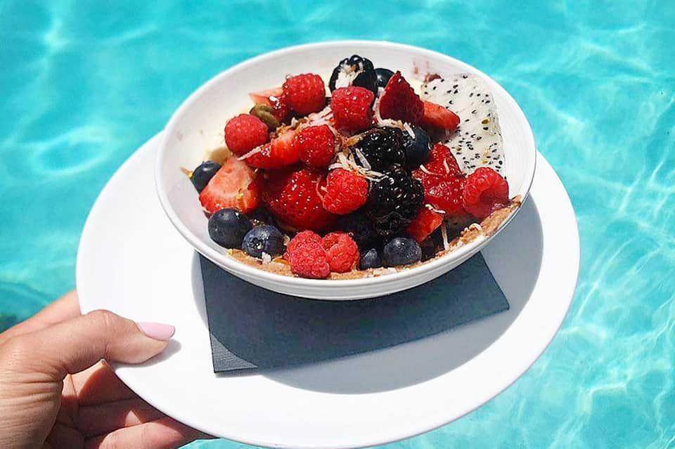 Bowl of granola with fresh raspberries, blueberries, and dragon fruit held over sparkling pool water