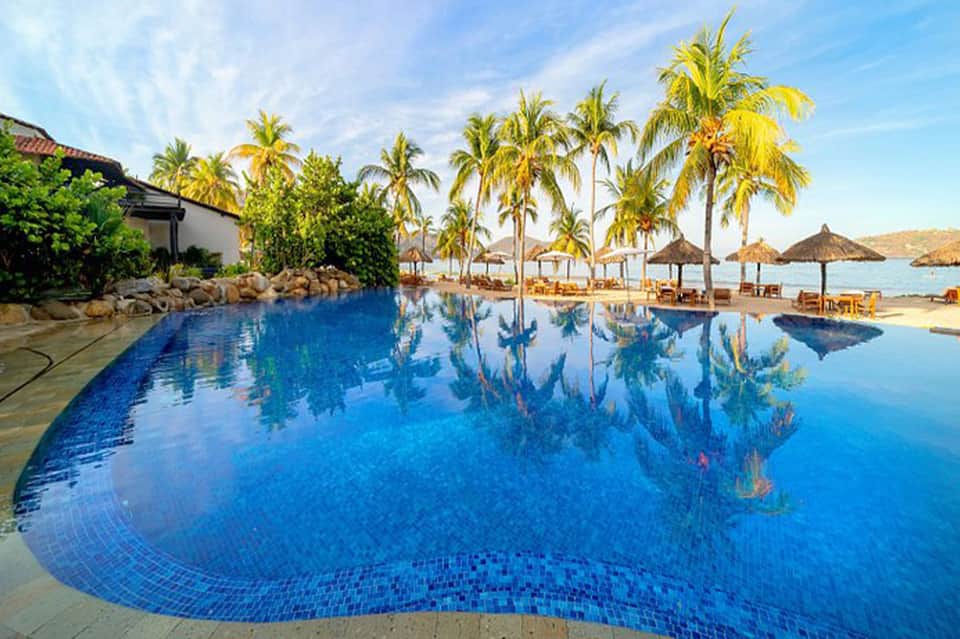 Infinity-edge resort pool with palm trees reflecting in still blue water, beach umbrellas and ocean view
