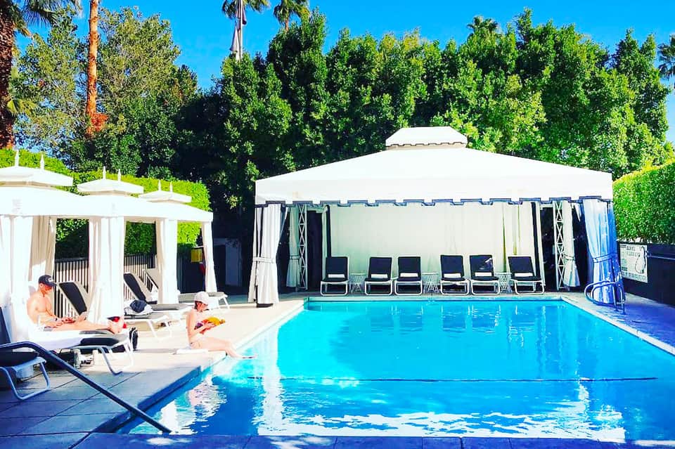 Resort pool with white cabana tent, lounge chairs, and lush green trees under clear blue sky