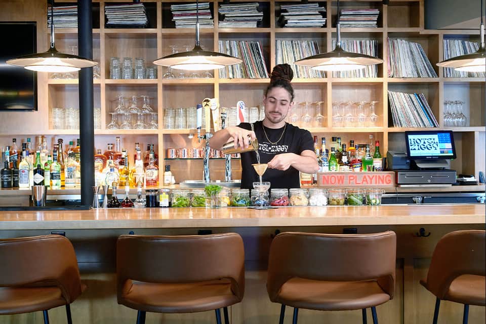 Craft cocktail bar with bartender mixing drinks, wooden shelving with spirits, and warm industrial lighting