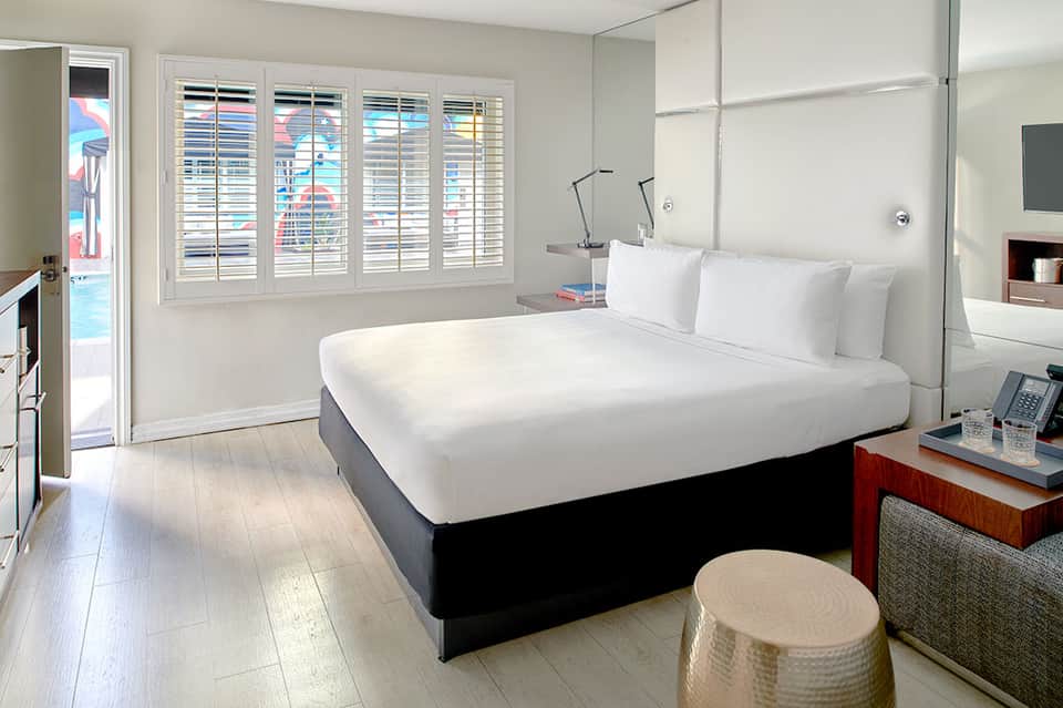 Modern hotel bedroom with white bedding, shuttered windows overlooking pool, and contemporary furnishings