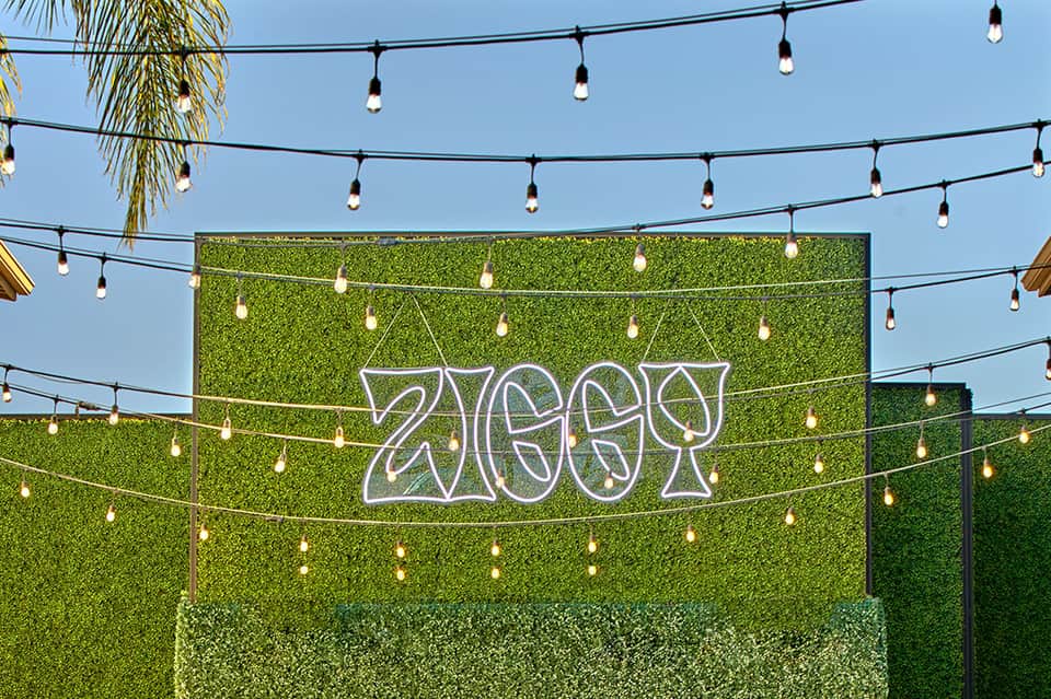 Outdoor event space with neon '2MGR' sign, green hedge walls, and hanging Edison bulb lighting