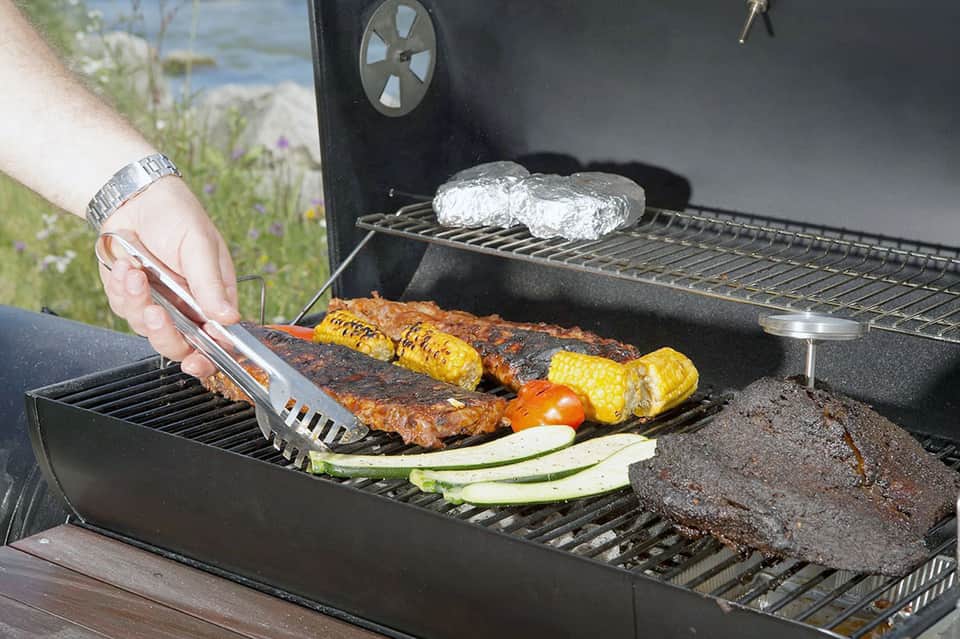 Person grilling ribs and vegetables on outdoor barbecue near water