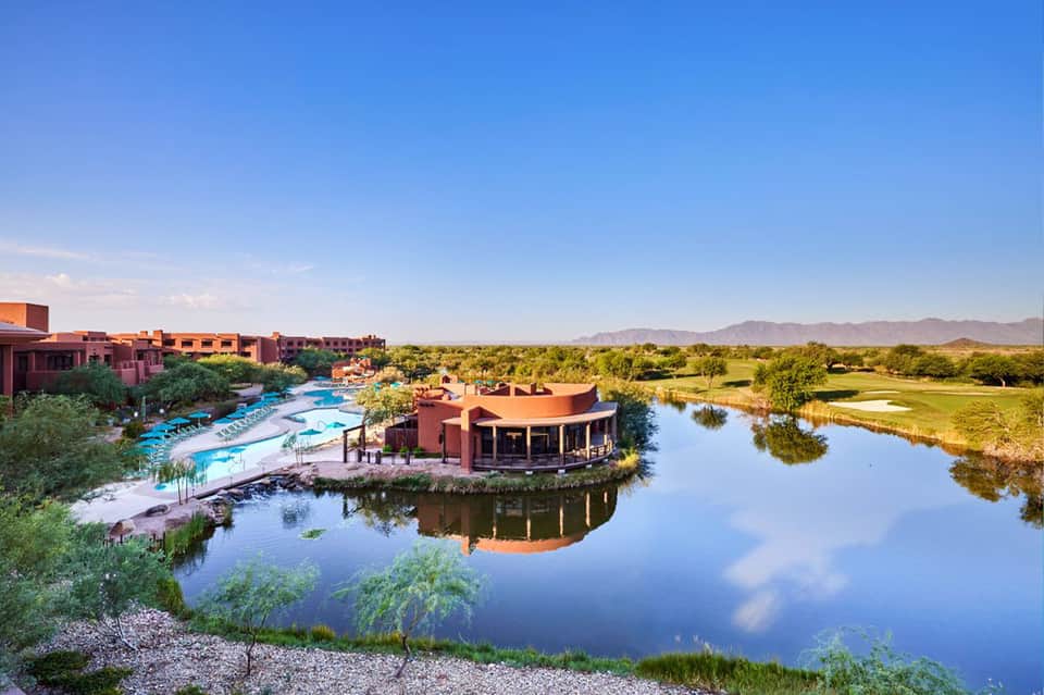 Aerial view of terracotta-colored resort buildings alongside a blue pool and waterway with landscaping