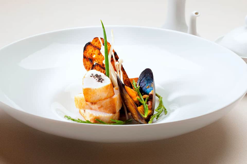 Elegant plated seafood dish with seared fish, mussel, and artistic garnish on white bowl
