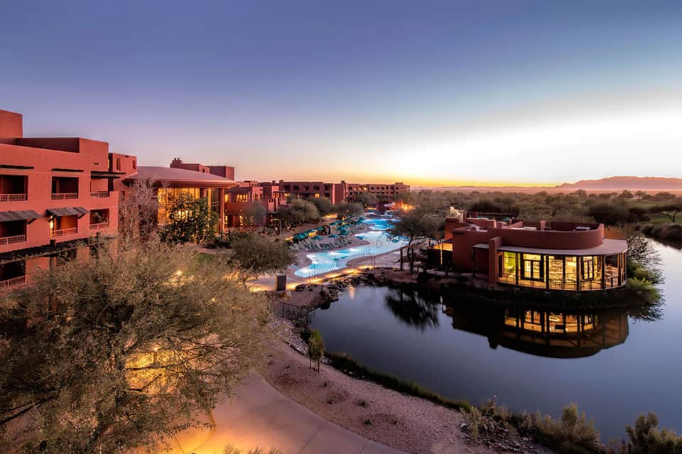 Red-clay resort buildings reflected in lagoon at dusk with illuminated pool, desert landscape, and evening sky