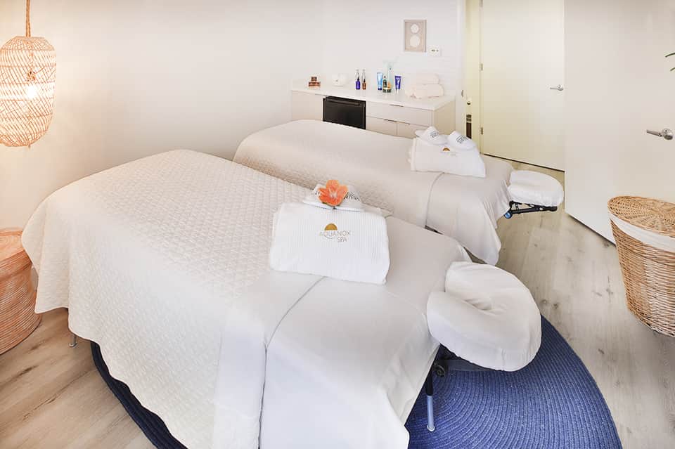 Spa treatment room with two massage tables and amenities counter with orange flower accent